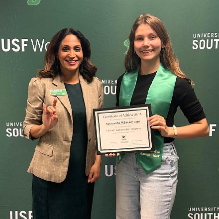Two people smiling and holding an award in front of a green background that says "USF World"