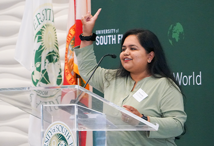 Dr. Nirmita Roy holding up hand with bulls sign and speaking at the podium