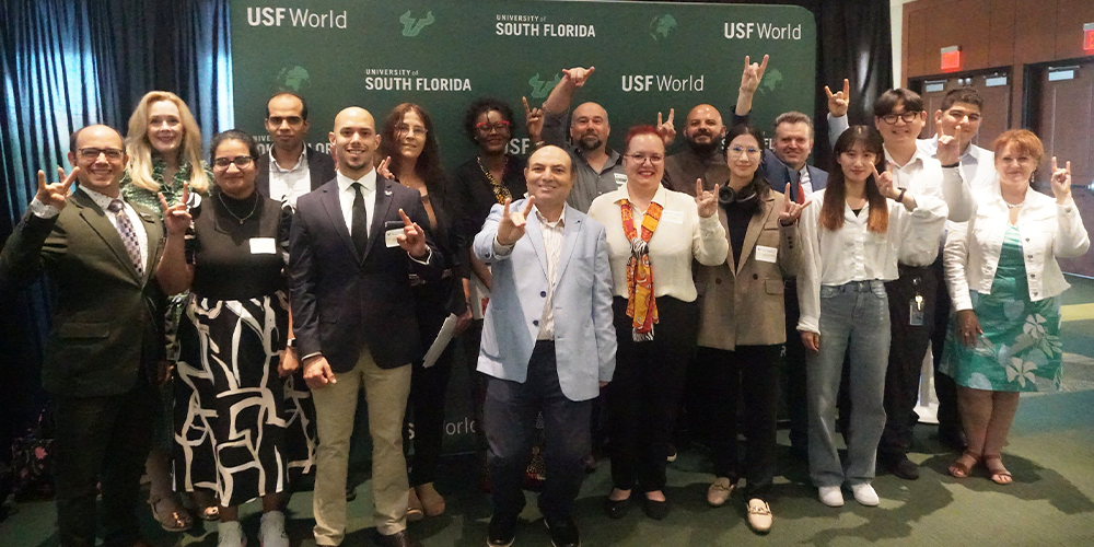 A group of smiling people holding up a bulls sign with their hands and standing in front of a green USF World backdrop