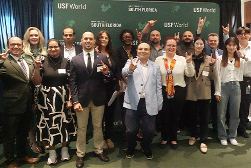 A group of smiling people holding up a bulls sign with their hands and standing in front of a green USF World backdrop