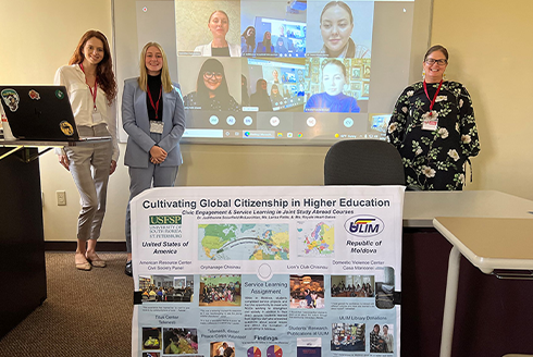 Professor and students in classroom standing behind a poster and beside a projector screen with students from around the world on a video call