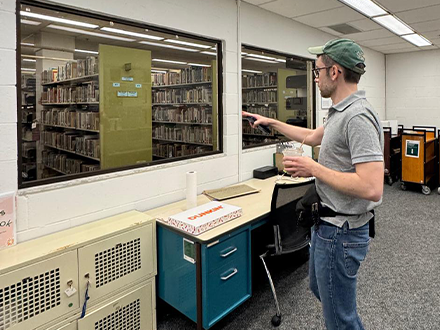 Carratt in the USF library pointing at the book on the shelves
