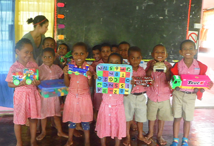 Group of children smiling and holding up play toys with adult in the background