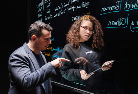 Two people looking at document on either side of a clear marker board