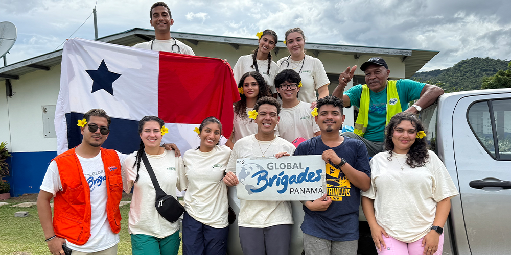 Large group of people standing on or below truck, someone holding a panama flag, while someone in the middle is holding a sign that says "Global Brigades"