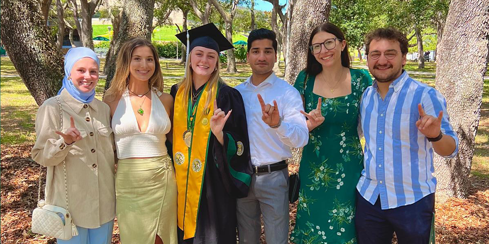 Taylor in a graduation stole and gown with a group of people smiling and holding up "go bulls" hand signs