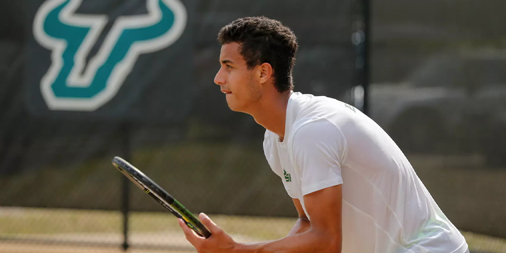 Roberto holding a tennis racket with the USF logo in the background