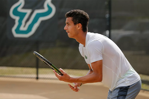 Roberto holding a tennis racket with the USF logo in the background