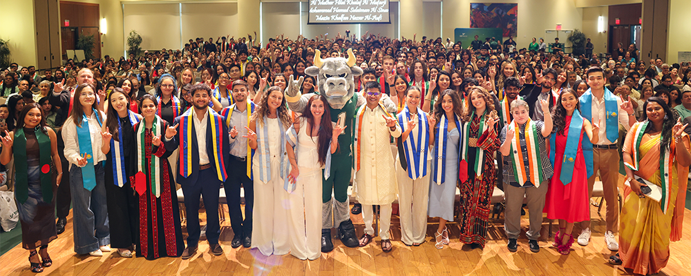 Group of international students with their flag stoles on, smiling and holding up go bulls signals with their hands. Rocky the Bull mascot is at the front.