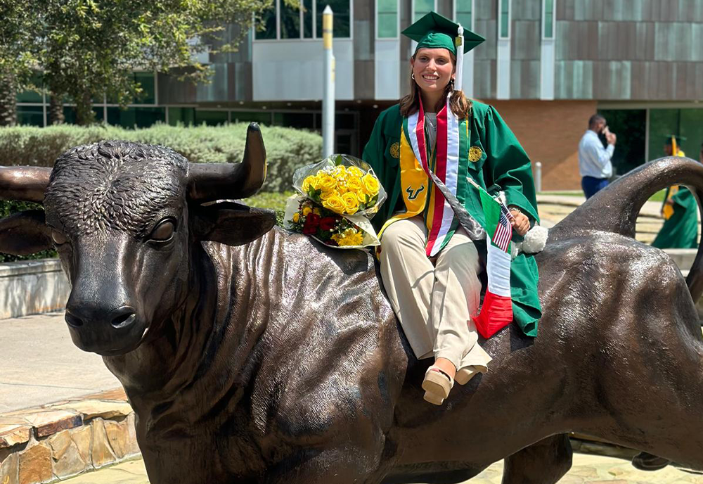 Student in sash, cap, and gown sitting on the USF Bull statue
