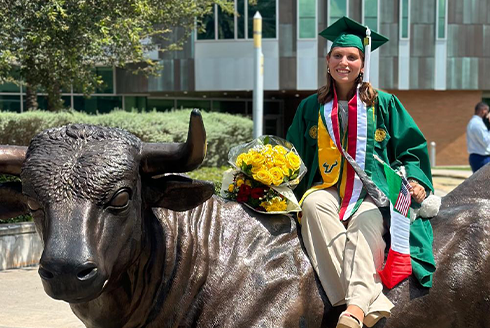 Student in sash, cap, and gown sitting on the USF Bull statue