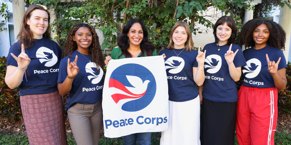 Six people stand with Peace Corps shirts on and the center person holding up a banner with the Peace Corps logo and text