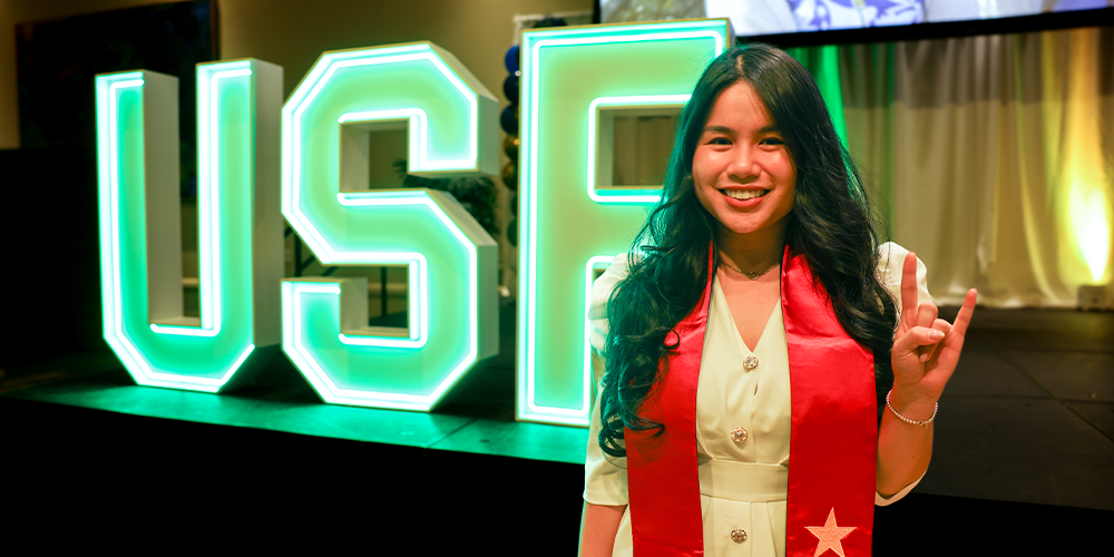 Bui smiling in front of a glowing neon "USF" sign